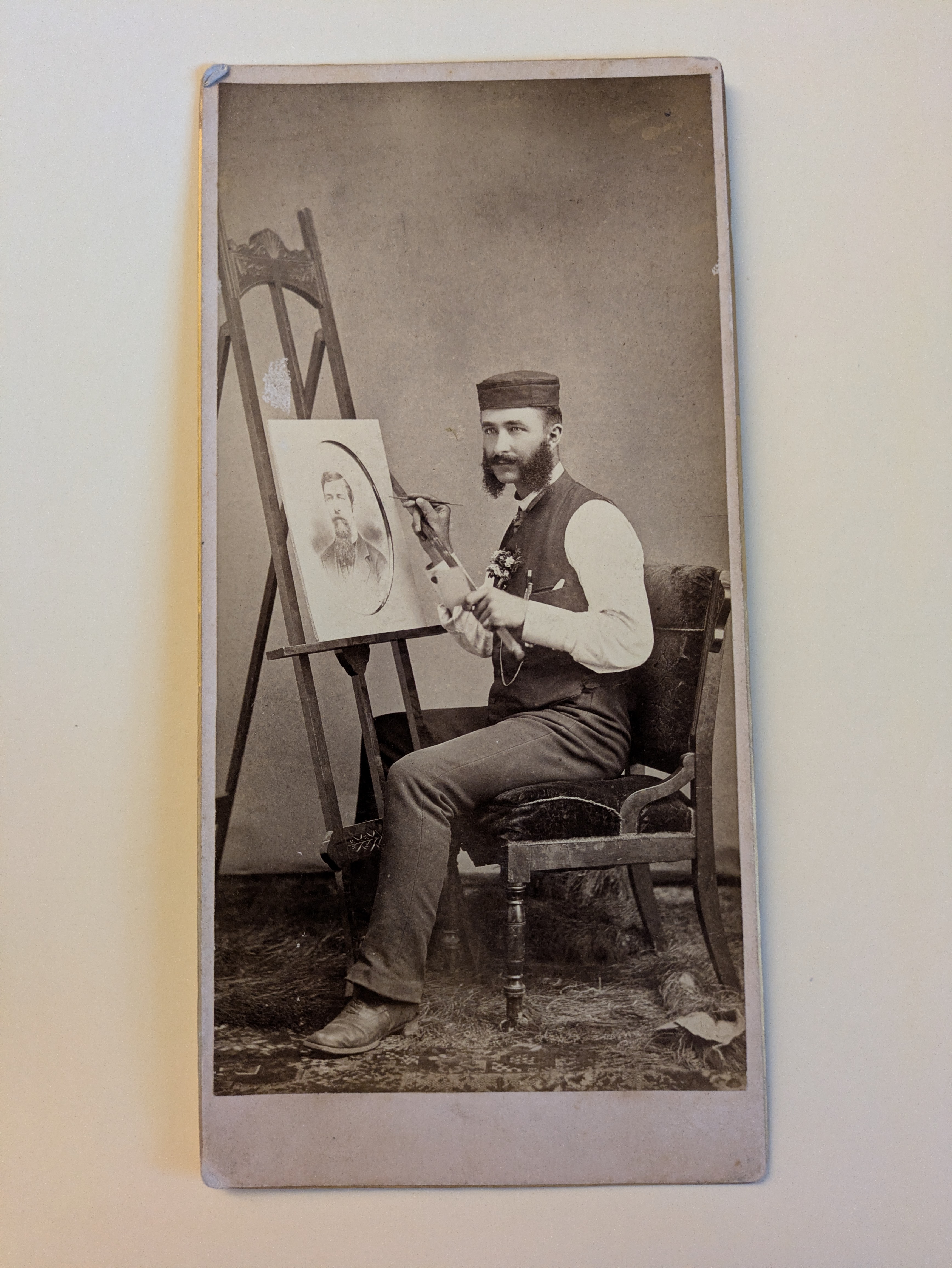 Artist Man seated on chair painting a portrait that is on an easel.