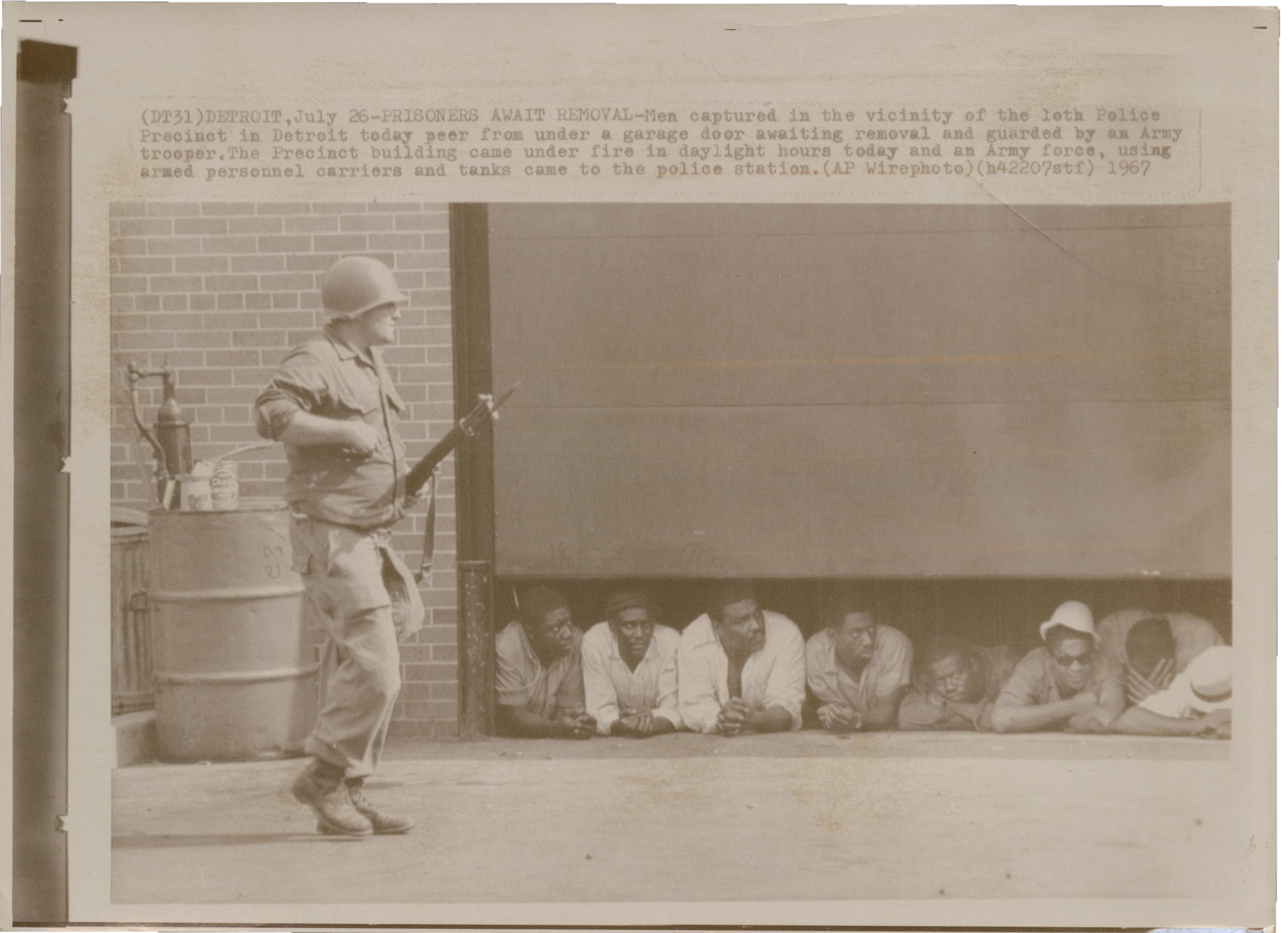 Detroit Riot A white man in fatigues wearing a helmet and carrying a large gun. Behind him, eight Black men lie on their stomachs under a lowered garage door. Typewritten text above the image describes the situation.