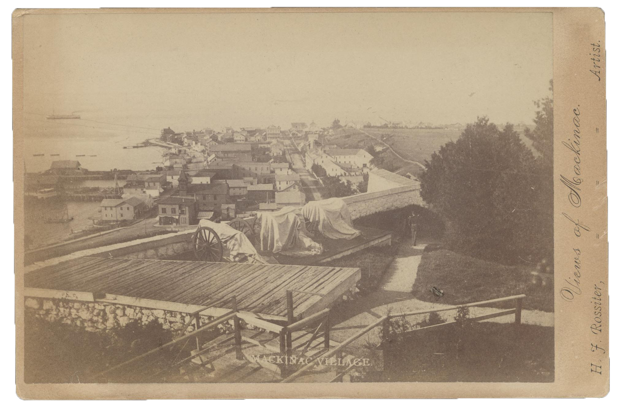 Mackinac Village Photograph of Mackinac Village from above, circa 1900. The town sits on the coast with several docks. In the foreground, cannons are covered by cloth.