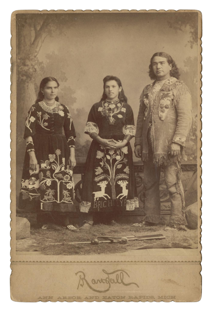 Susette La Flesche, Francis La Flesche, and a Ponca Sioux Mother - Edited Full body photograph of Susette La Flesche, Francis La Flesche, and a Ponca Sioux Mother. Their clothes are intricate, with sewn-on depictions of animals and flowers. Susette La Flesche has the name “Bright Eye” on the bottom of her dress in large letters.